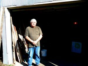 man with white hair and white mustache standing before the gaping entrance of a warehouse-like workshop