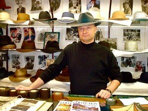 Gary Witkowski, custom hatter, at his shop, with rolls of ribbon on the table and hats behind him